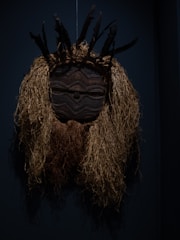 A tribal mask made of dark wood with intricate carvings, featuring eye patterns and a central diagonal stripe. The mask is adorned with a headdress of black feathers and long straw-like material that resembles hair.