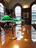 A long wooden table is lined with vintage brass lamps featuring green glass shades, situated in a room with tall arched windows and rich wood paneling. A portrait is hung on the wall, and there are wooden chairs along the sides of the table.