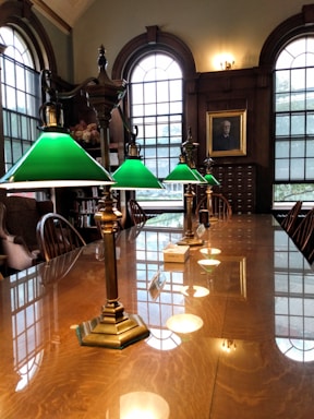 A long wooden table is lined with vintage brass lamps featuring green glass shades, situated in a room with tall arched windows and rich wood paneling. A portrait is hung on the wall, and there are wooden chairs along the sides of the table.