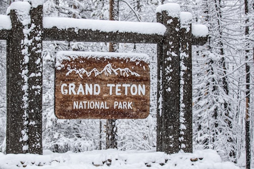 A wooden sign covered in snow with the words 'Grand Teton National Park' carved into it. The sign hangs from a larger wooden structure that is also dusted with snow. In the background, snow-covered trees create a wintery forest scene.