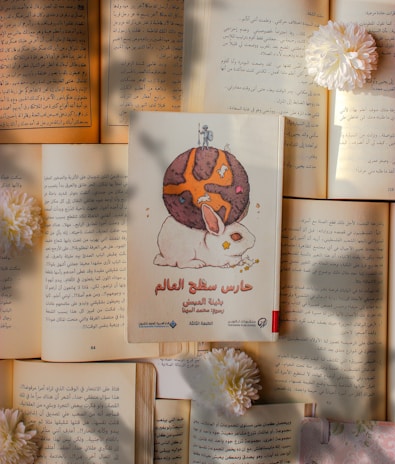 A book with an illustration of a rabbit and a brown planet or sphere sits atop an array of opened books. The pages feature text in Arabic script, and several white chrysanthemum flowers are placed around the arrangement, casting gentle shadows.