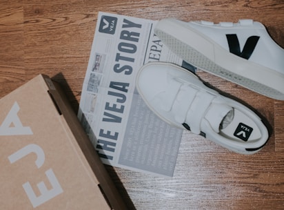 A pair of white sneakers with a black 'V' logo is placed on a wooden floor alongside a cardboard shoe box and a newspaper-style brochure. The brochure features text about 'The Veja Story' and includes some images.