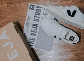 A pair of white sneakers with a black 'V' logo is placed on a wooden floor alongside a cardboard shoe box and a newspaper-style brochure. The brochure features text about 'The Veja Story' and includes some images.