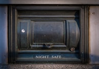 A secure metal box embedded into the wall, labeled 'Night Safe.' It has a sturdy and slightly worn appearance, with evident use and weathering.