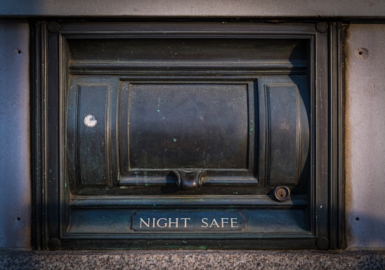 A secure metal box embedded into the wall, labeled 'Night Safe.' It has a sturdy and slightly worn appearance, with evident use and weathering.