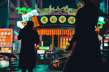 Bright neon lights illuminate a bustling street market adorned with vibrant signage and decorative lanterns. Silhouettes of people in the foreground add depth and a sense of busy activity. The scene captures the essence of a lively urban night environment.