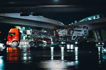 A large red truck is shown transporting heavy machinery on a low loader trailer under an overpass at night. The scene reflects on the wet road surface beneath, suggesting recent rain. The road and overpass are illuminated with artificial lights, casting reflections in the water.
