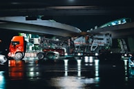 A large red truck is shown transporting heavy machinery on a low loader trailer under an overpass at night. The scene reflects on the wet road surface beneath, suggesting recent rain. The road and overpass are illuminated with artificial lights, casting reflections in the water.