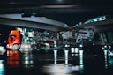 A large red truck is shown transporting heavy machinery on a low loader trailer under an overpass at night. The scene reflects on the wet road surface beneath, suggesting recent rain. The road and overpass are illuminated with artificial lights, casting reflections in the water.