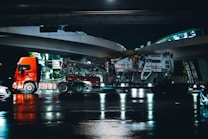 A large red truck is shown transporting heavy machinery on a low loader trailer under an overpass at night. The scene reflects on the wet road surface beneath, suggesting recent rain. The road and overpass are illuminated with artificial lights, casting reflections in the water.