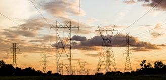 A landscape featuring multiple high-voltage power lines stretching across a field with a dramatic sky. The sun is setting, casting warm hues across scattered clouds and silhouetting the steel towers.