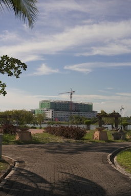 A paved path leads towards a large building under construction, surrounded by greenery. A crane is visible above the building, which is partially covered with green netting. The scene is set against a backdrop of a blue sky with white clouds. Trees and neatly trimmed bushes line the area, contributing to a tranquil environment. Palm fronds and the shadow of lamp posts appear in the foreground.