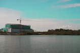 A construction site with a multi-story building partially covered in green netting is visible by the waterfront. A tall crane is positioned on top of the building, extending over the structure. The scene is set against a backdrop of trees and a partly cloudy sky with a turquoise tint in the upper portion.