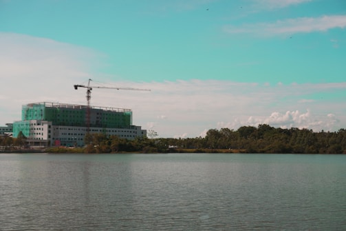 A construction site with a multi-story building partially covered in green netting is visible by the waterfront. A tall crane is positioned on top of the building, extending over the structure. The scene is set against a backdrop of trees and a partly cloudy sky with a turquoise tint in the upper portion.