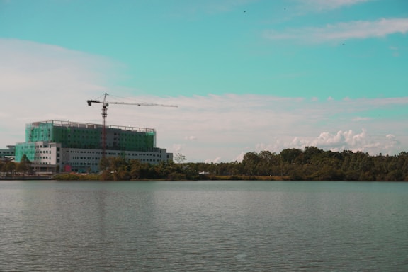 A construction site with a multi-story building partially covered in green netting is visible by the waterfront. A tall crane is positioned on top of the building, extending over the structure. The scene is set against a backdrop of trees and a partly cloudy sky with a turquoise tint in the upper portion.