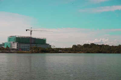 A construction site with a multi-story building partially covered in green netting is visible by the waterfront. A tall crane is positioned on top of the building, extending over the structure. The scene is set against a backdrop of trees and a partly cloudy sky with a turquoise tint in the upper portion.