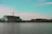 A construction site with a multi-story building partially covered in green netting is visible by the waterfront. A tall crane is positioned on top of the building, extending over the structure. The scene is set against a backdrop of trees and a partly cloudy sky with a turquoise tint in the upper portion.