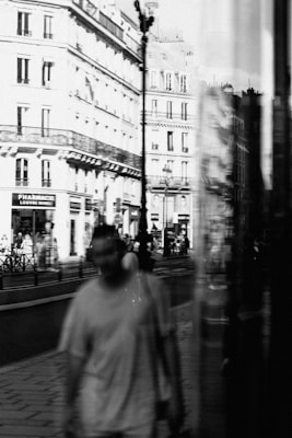 A black and white photograph depicts an urban street scene. There is a reflection in a glass window of a person walking and a view of historic buildings. The scene captures pedestrians and a pharmacy with the name Louvre visible. The atmosphere feels bustling and lively.