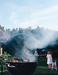A barbecue grill with smoke rising from it is set in a lush green garden. There are children in the background, one with a stroller, walking on the grass. Trees and a trampoline are visible in the distance under a clear blue sky.