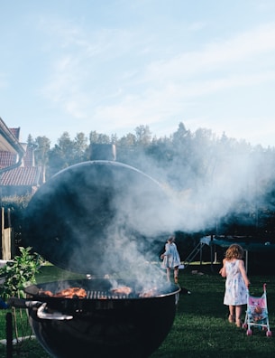 A barbecue grill with smoke rising from it is set in a lush green garden. There are children in the background, one with a stroller, walking on the grass. Trees and a trampoline are visible in the distance under a clear blue sky.
