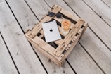 A metallic tablet with a recognizable logo is placed on a rustic wooden crate, alongside a pair of sunglasses and a tall glass of iced coffee. The setting is outdoors on a wooden deck, creating a relaxed and casual atmosphere.