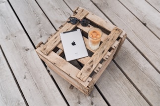 A metallic tablet with a recognizable logo is placed on a rustic wooden crate, alongside a pair of sunglasses and a tall glass of iced coffee. The setting is outdoors on a wooden deck, creating a relaxed and casual atmosphere.