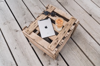 A metallic tablet with a recognizable logo is placed on a rustic wooden crate, alongside a pair of sunglasses and a tall glass of iced coffee. The setting is outdoors on a wooden deck, creating a relaxed and casual atmosphere.