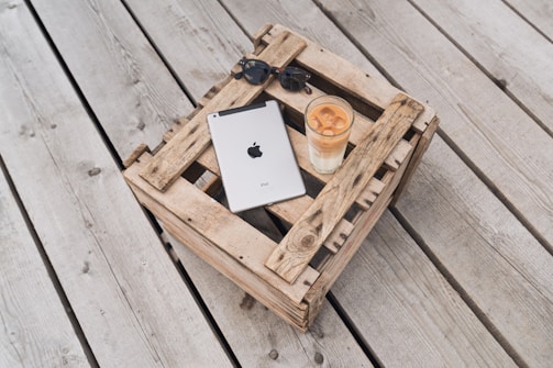 A metallic tablet with a recognizable logo is placed on a rustic wooden crate, alongside a pair of sunglasses and a tall glass of iced coffee. The setting is outdoors on a wooden deck, creating a relaxed and casual atmosphere.
