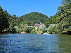 A serene landscape featuring a large body of water surrounded by lush greenery and dense forest. A charming building with a red-tiled roof is nestled among the trees in the background, with rolling hills rising behind it under a clear blue sky.