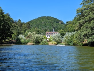 A serene landscape featuring a large body of water surrounded by lush greenery and dense forest. A charming building with a red-tiled roof is nestled among the trees in the background, with rolling hills rising behind it under a clear blue sky.