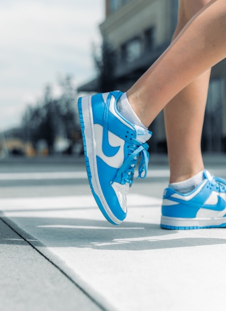 Blue and white sneakers are shown in mid-air, worn by a person on a sunlit sidewalk. The background is slightly blurred, indicating an urban setting.