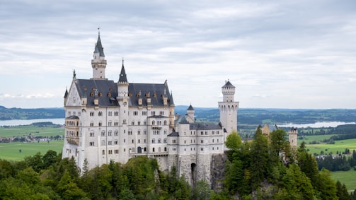 A large, historic castle with multiple towers and spires set atop a lush, forested hill. The building is constructed with light-colored stone and features ornate architectural designs. The background shows a vast, scenic landscape with rolling hills and distant water bodies under a cloudy sky.