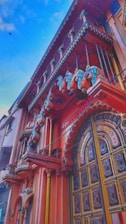 A vibrant architectural facade featuring intricate designs and colorful embellishments. The building showcases ornate balconies and a large, decorated wooden door with detailed carvings. The architecture exudes a traditional style with vivid colors and artistic elements including decorative sculptures.