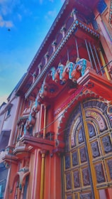 A vibrant architectural facade featuring intricate designs and colorful embellishments. The building showcases ornate balconies and a large, decorated wooden door with detailed carvings. The architecture exudes a traditional style with vivid colors and artistic elements including decorative sculptures.