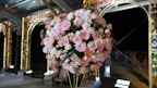 A large, ornate floral arrangement featuring an assortment of pink and white roses along with some peach-colored flowers. The flowers are intricately arranged into a spherical shape and situated in a lavish, illuminated outdoor setting. Elegant arches and decorative lights provide a backdrop, enhancing the sophisticated atmosphere.