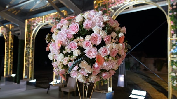 A large, ornate floral arrangement featuring an assortment of pink and white roses along with some peach-colored flowers. The flowers are intricately arranged into a spherical shape and situated in a lavish, illuminated outdoor setting. Elegant arches and decorative lights provide a backdrop, enhancing the sophisticated atmosphere.