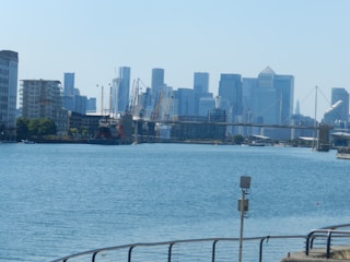 A cityscape with a skyline of modern skyscrapers along the waterfront. A body of water is in the foreground, with a metal railing and security camera in view. Construction cranes are visible in the distance, and a bridge crosses the water.