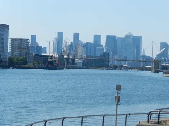 A cityscape with a skyline of modern skyscrapers along the waterfront. A body of water is in the foreground, with a metal railing and security camera in view. Construction cranes are visible in the distance, and a bridge crosses the water.