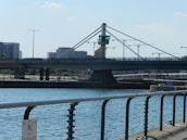 A modern bridge with cables and a central tower spans across a body of water. In the background, several high-rise buildings and crane structures indicate construction activities. The scene includes elements of urban infrastructure, like street lamps and railings along the waterfront.