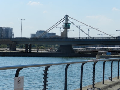 A modern bridge with cables and a central tower spans across a body of water. In the background, several high-rise buildings and crane structures indicate construction activities. The scene includes elements of urban infrastructure, like street lamps and railings along the waterfront.