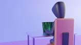 A 3D-rendered figure sits at a table, typing on a laptop with green code on the screen. The environment is minimalist and features soft lighting with a pastel color scheme.