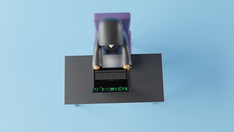 A 3D-rendered character is sitting at a desk using a laptop. The character appears simplified and cartoon-like, viewed from above. The desk is dark-colored with a laptop showing a grid and green digits, suggesting a futuristic or digital theme.