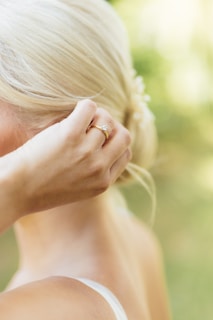 A close-up of a person with blonde hair styled in an elegant updo. The focus is on their hand, adorned with a delicate gold ring featuring a small gemstone. The soft background suggests a natural, outdoor setting with lush greenery.