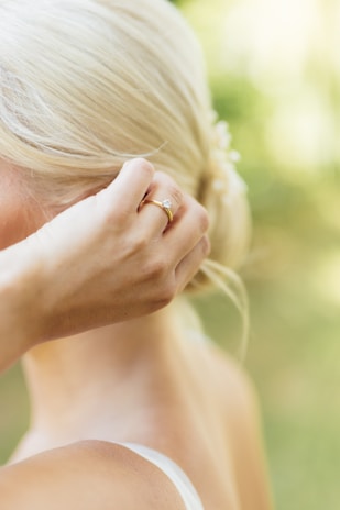 A close-up of a person with blonde hair styled in an elegant updo. The focus is on their hand, adorned with a delicate gold ring featuring a small gemstone. The soft background suggests a natural, outdoor setting with lush greenery.