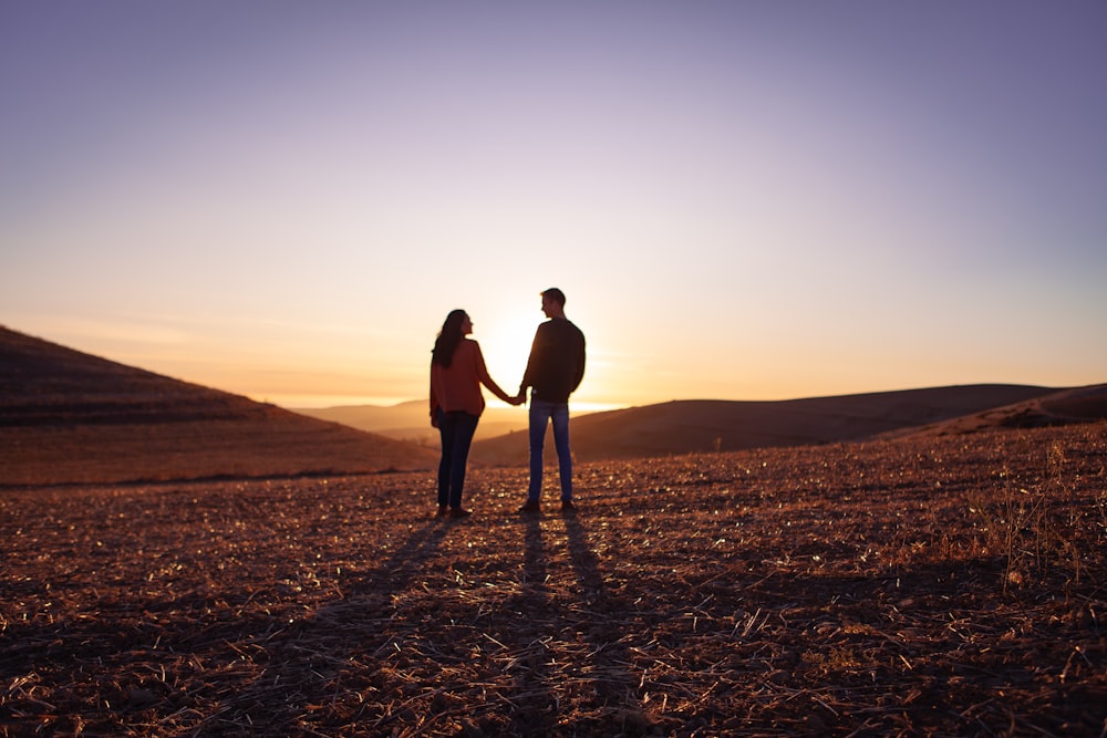 a man and woman standing on a dirt road with the sun setting