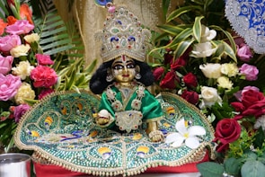 A beautifully adorned religious idol placed on an ornate, green, jeweled cloth. The idol has elaborate decorations, including a large, colorful crown and intricate clothing. It is surrounded by an assortment of vibrant flowers, including roses and lilies, with green foliage in the background.