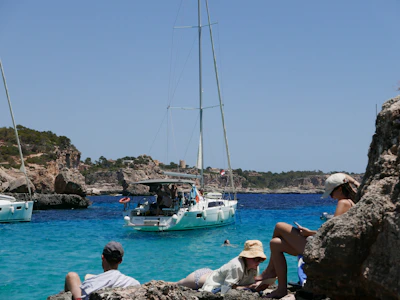 people sitting on rocks looking at a sailboat in the water