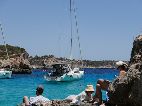 people sitting on rocks looking at a sailboat in the water