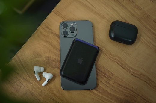 A smartphone with a black wallet attached to its back is placed on a wooden surface. Next to the smartphone are a pair of white wireless earbuds and a black carrying case. There is a hint of green foliage in the top left corner.