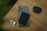 A smartphone with a black wallet attached to its back is placed on a wooden surface. Next to the smartphone are a pair of white wireless earbuds and a black carrying case. There is a hint of green foliage in the top left corner.
